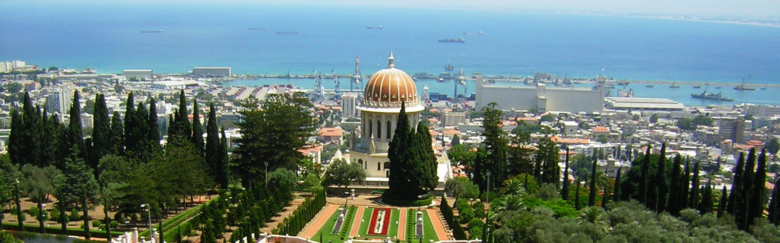 A circular, white building with arches has a golden dome, surrounded by a city and a large body of water in the distance. 